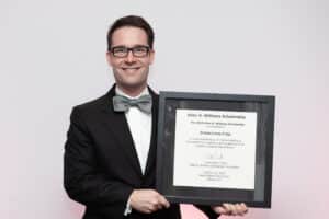 A man in a bow tie and suit stands smiling, holding a framed certificate titled "Allen D. Williams Scholarship" awarded to "Jonathan Lewis, P. Eng.", for contributions to the consulting engineering industry, alongside his recent accolade: the prestigious Graham Lovely Award.