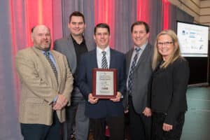 Five individuals are standing in front of a gray and red backdrop at the UNB Fredericton Kinesiology Building, with one person in the center holding an ACEC Pinnacle Award plaque. They are all smiling and dressed in formal attire.