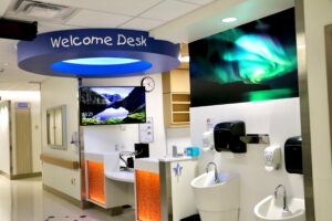 A welcome desk in a modern building with a curved blue sign overhead. The desk has a computer, clock, and landscape screensaver. Nearby are sinks and soap dispensers beneath a photo of the Northern Lights, embodying the spirit of Stollery Children’s Hospital's Pediatric Critical Care unit.