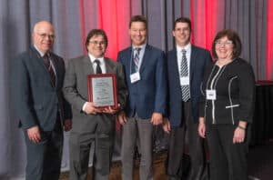 A group of five people in business attire stands together smiling, with one person holding an ACEC Engineering Award plaque. Red and gray curtains serve as the backdrop.