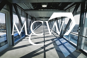 A modern glass corridor under a steel framework with sunlight streaming through windows. The bold letters "MCW Transportation" overlay the image, highlighting this impressive transportation feature.
