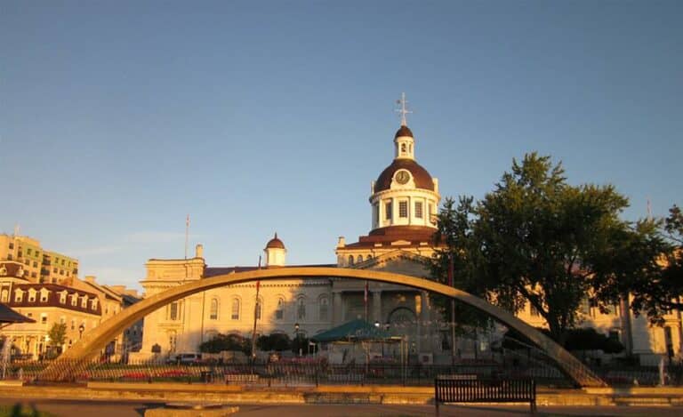 Historic building with a central dome and clock tower, flanked by trees and smaller turrets, viewed behind a modern arched structure in front along with benches and a park area at sunset in the City of Kingston.