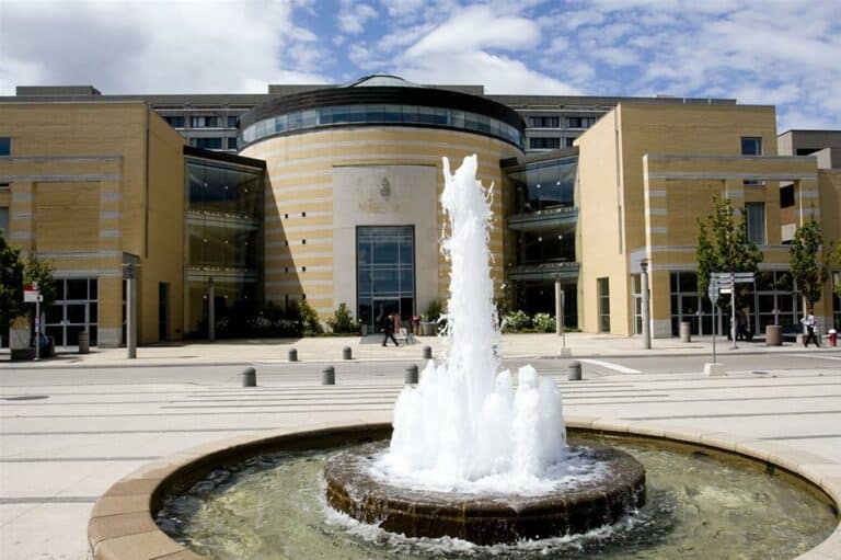 Modern building with a circular main entrance and glass dome, flanked by symmetrical wings, featuring a fountain in the foreground. The sky is partly cloudy. Located within York University’s campus, this structure embodies both innovation and tradition, creating an inspiring academic environment.