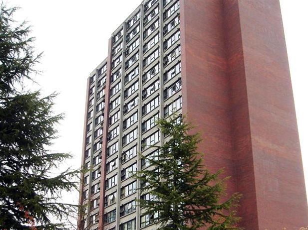 A tall brick apartment building with multiple windows, part of BC Housing, is surrounded by a few evergreen trees under a cloudy sky.