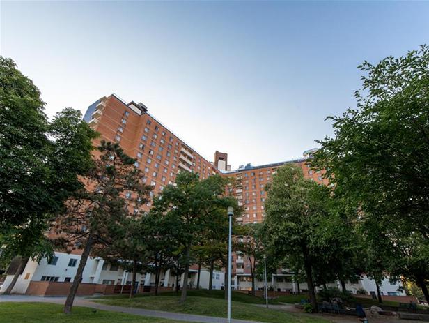 A large, multi-story brick apartment building, part of Toronto Community Housing, is surrounded by trees on a clear day. Pathways and lampposts are visible in the foreground.