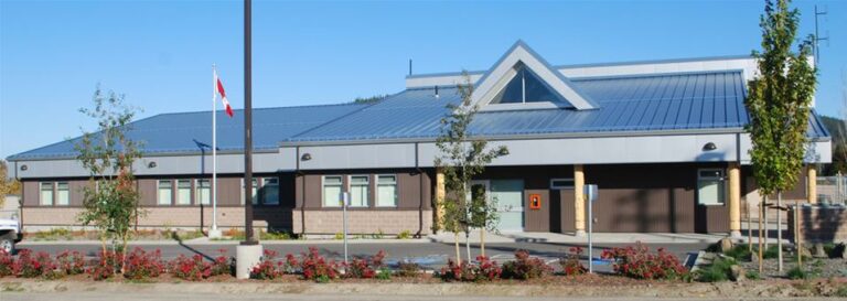 Single-story building with a blue metal roof, several small trees and plants in front, and a flagpole with a Canadian flag on the left side. The building features large windows and a triangular roof peak, giving it an official look reminiscent of an RCMP station.