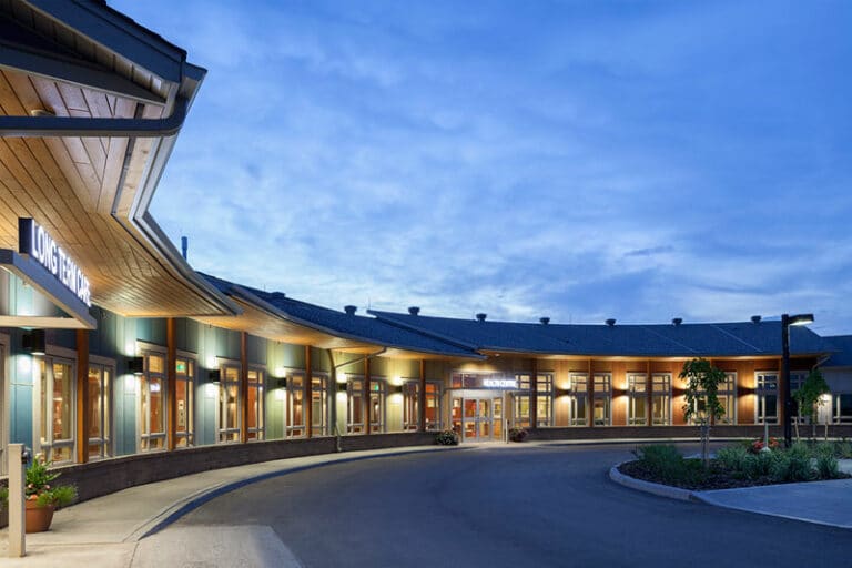 A curved building with numerous windows and a central entrance is illuminated at dusk. The sign at the entrance reads "URGENT PRIMARY CARE CENTRE," a vital facility within the Sun Country Health Region. A circular driveway surrounds the entrance, ensuring convenient access for all.