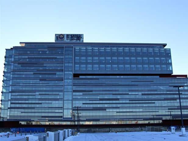 A modern multi-story office building with extensive glass windows, labeled "Regado Biosciences" at the top. In the foreground, construction materials and equipment hint at ongoing development. The sky is clear and blue. Nearby, ties to the University of Alberta signal innovation and collaboration in progress.