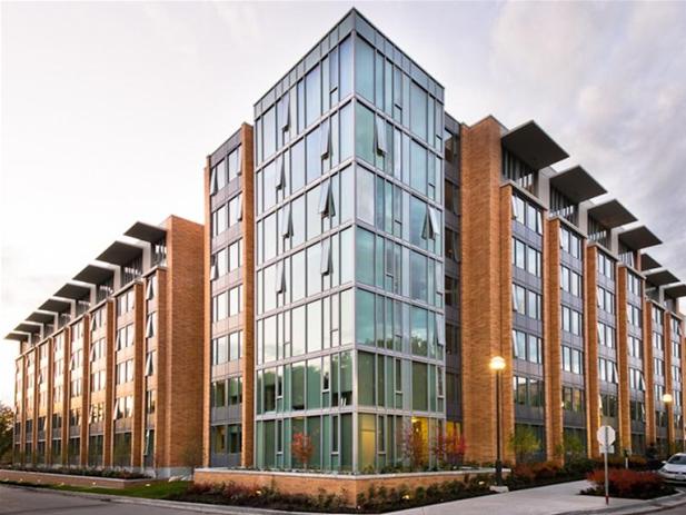 A modern multi-story building with large glass windows and brick accents, set against a partly cloudy sky with lush landscaping in the foreground, reminiscent of the architectural style found at UBC.