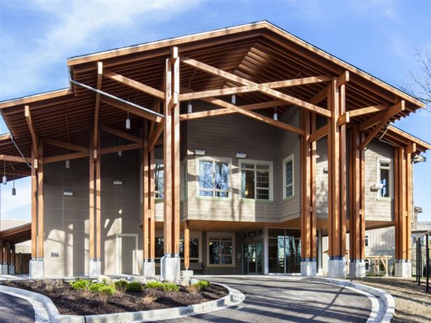 A modern building in British Columbia features large wooden beams and slanted roofs, with extensive windows on the ground and first floors; a paved driveway leads to the entrance. This facility, part of Fraser Health, exemplifies cutting-edge health care infrastructure.