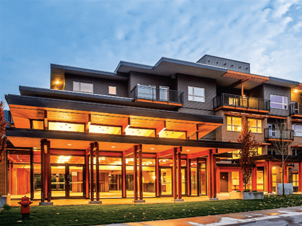 A modern, multi-story building with large glass windows and balconies, illuminated from inside, sits elegantly under a twilight sky in the serene neighborhood of West Vancouver's Kiwanis Housing Society.