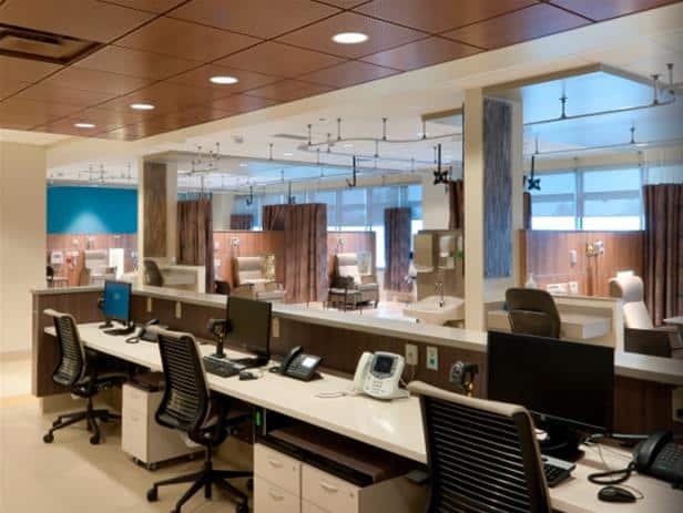 A modern hospital workstation area with multiple computers, chairs, and phones provided by Alberta Health Services. In the background, there are patient bays separated by curtains. The room is well-lit with overhead lighting.