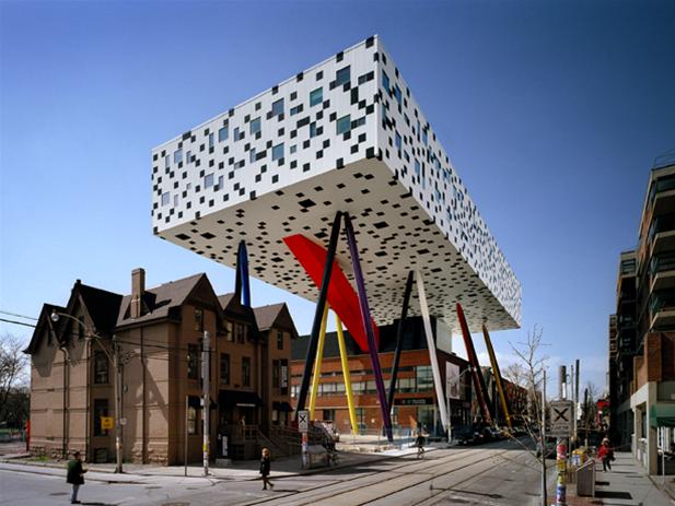 A modern building with a large pixelated box design rests on multicolored stilts above traditional houses on a city street, embodying the innovative spirit of the Ontario College of Art and Design.