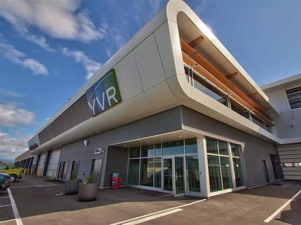 Modern commercial building with large glass windows and an upper-level terrace, featuring the initials "VR" on the facade. Situated under a blue sky with clouds, it's conveniently located near Vancouver International Airport.