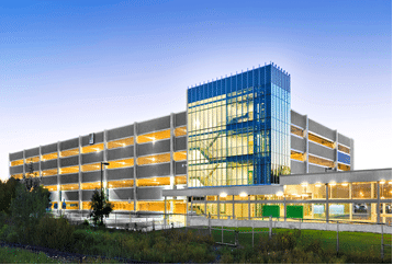 A multi-story parking structure with an illuminated stairwell at dusk, featuring open sides and surrounded by greenery, designed in collaboration with Metrolinx for seamless integration with GO Transit services.