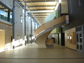 A modern indoor atrium with high ceilings, featuring a curved, wooden staircase and natural light streaming through upper windows, is an architectural highlight often seen in the Trillium Lakehead District School Board.