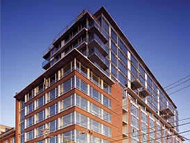 A modern multi-story building with large glass windows and brick accents is seen from a low angle against a clear blue sky, showcasing the innovative design hallmark of Context Developments Inc.