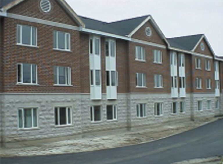 A three-story brick apartment building with pitched roofs and multiple windows stands majestically nearby a bustling college campus. The lower section of the building is constructed from light-colored stone. A newly paved road runs in front, adding to its charming appeal.