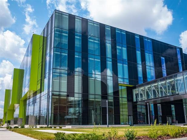 Modern glass building with green accents, featuring reflective windows and a connected walkway. Blue sky with clouds in the background and a landscaped green area in the foreground, perfectly encapsulating the vibrant campus life of MacEwan University.