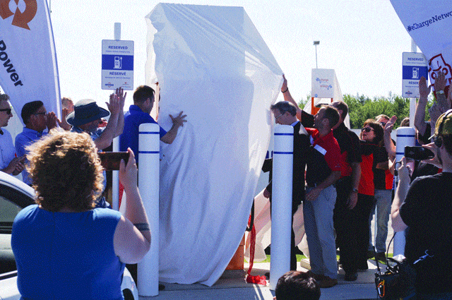 A group of people gathers around and unveils a large covered object at an outdoor event in New Brunswick, with some onlookers taking photos and holding signs. The event highlights MCW Supports’ commitment to innovation, featuring a display of cutting-edge battery-powered driving technology.